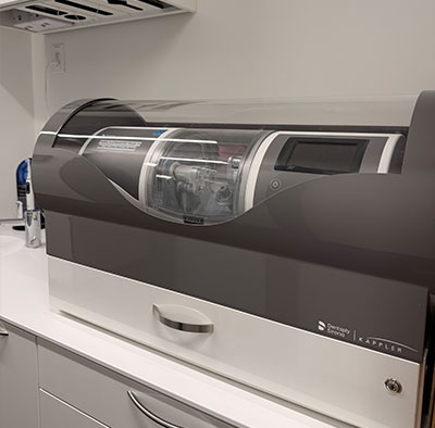 The image shows a modern-looking appliance with a clear glass door, likely a laboratory equipment or a machine for scientific research, placed on a countertop next to a white wall.