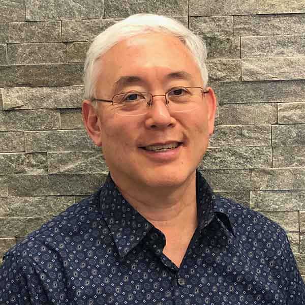 The image shows a man smiling at the camera. He has short hair, wears glasses, and is dressed in a patterned shirt with a blue collar. In the background, there s a wall of gray tiles.