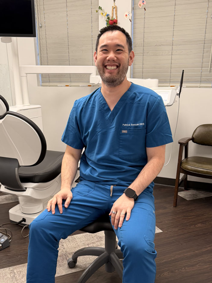 The image shows a smiling man wearing blue scrubs, sitting on a chair with his hands clasped together in front of him, likely in an office or dental practice setting.