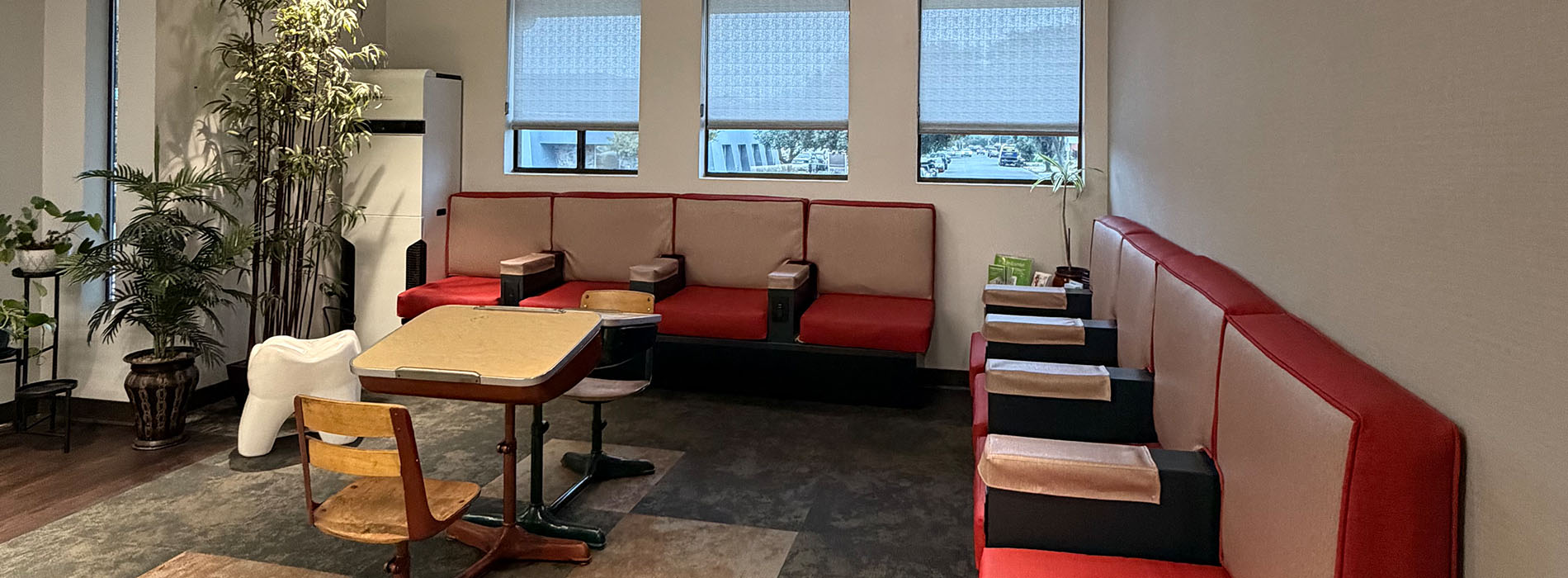 This is a photograph of an interior space featuring several dining tables with red benches, a wall-mounted TV, and decorative elements such as potted plants and framed artwork. The room has a modern aesthetic with clean lines and minimalist design.