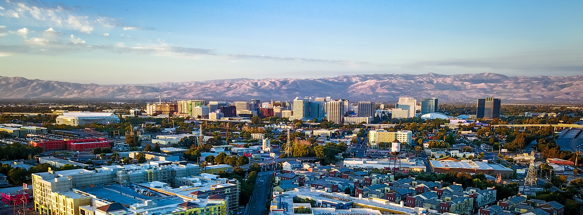 The image shows a panoramic view of an urban landscape with a city skyline under a clear blue sky during sunset, featuring various buildings, including a prominent tower, against a backdrop of mountains.