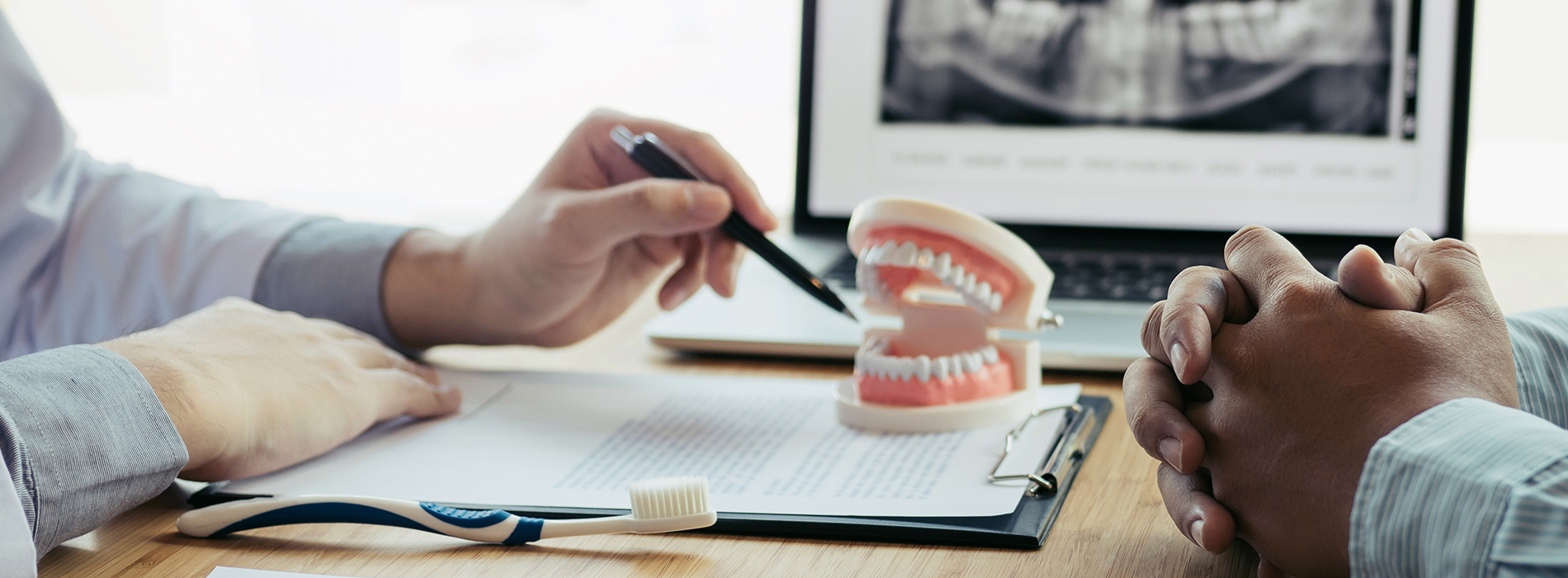 This is a photograph featuring two individuals seated at a table with documents one person appears to be a dental professional examining an open mouth model, while the other individual is recording or taking notes on a tablet. The setting suggests a dental consultation or examination room, and there are medical models and equipment visible in the background.