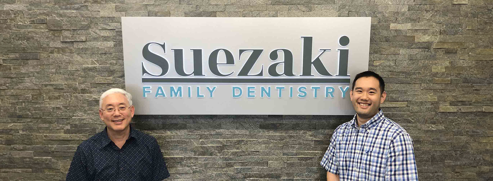 The image shows two individuals standing in front of a sign for Sugaki Family Dentistry, with a backdrop featuring a brick wall and a logo.