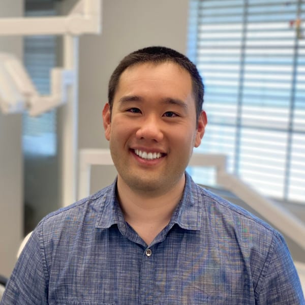 The image shows a man smiling at the camera, standing in front of dental equipment with a bright background.