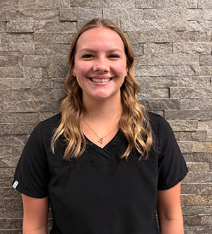 A woman wearing a black shirt with a name tag, smiling at the camera while standing against a brick wall.