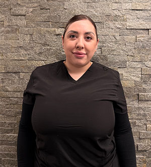 The image shows a woman standing against a brick wall with her arms crossed, wearing a black shirt and dark pants, and has short hair.