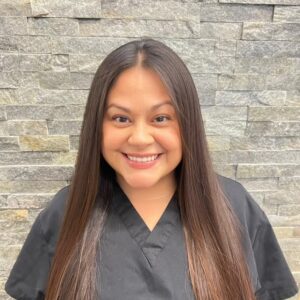 The image shows a person smiling at the camera, wearing a black shirt with a collar, standing in front of a stone wall with a light-colored background.