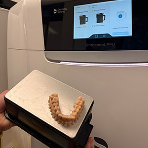 The image shows a person holding up a tooth with a 3D printer in the background, displaying the process of 3D printing teeth.