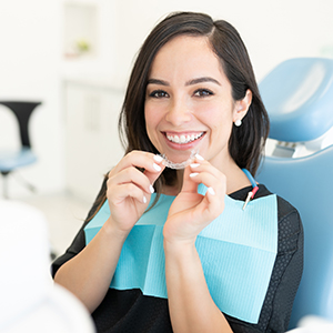 The image shows a woman sitting at a dental chair with a smile, holding dental tools, and wearing a blue face mask.