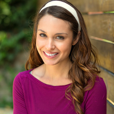 A young woman with long brown hair wearing a purple top and a white headband is smiling at the camera.