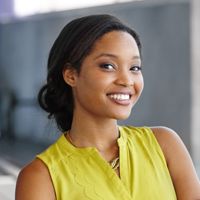 The image shows a smiling woman with short hair wearing a yellow top and standing against a backdrop with a modern architectural design, possibly a building facade.