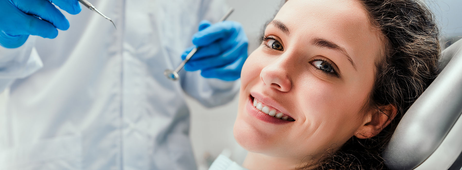 The image shows a woman with a smiling expression, wearing blue gloves and sitting in front of dental equipment, possibly in a dental office setting.