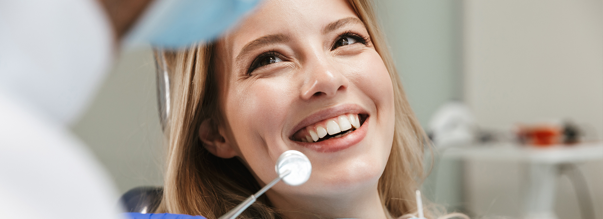 The image features a smiling woman with blonde hair sitting in a dental chair, receiving dental care, with a dental professional standing nearby holding a mirror.