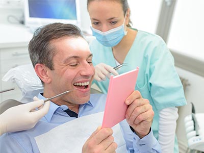 Man sitting in dental chair with paper over his mouth, surrounded by dental professionals  he appears surprised or shocked.