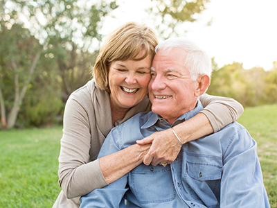 The image shows two older adults, an elderly man and woman, embracing each other with smiles on their faces while sitting outdoors during daylight.