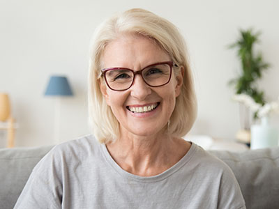 A woman with short hair is smiling at the camera, wearing glasses and a casual top. She appears to be indoors, possibly in a living room setting.
