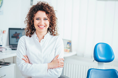 The image shows a woman standing in an office environment, wearing a white shirt, smiling at the camera with her hands on her hips.