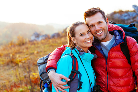 A man and woman are standing close together outdoors, with the man wearing a backpack, both smiling at the camera, during daylight.