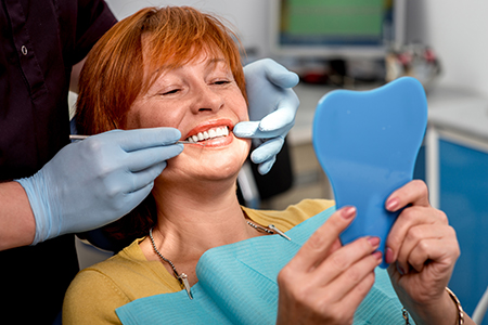 The image shows a woman sitting in a dental chair with a toothbrush-shaped object placed on her teeth, looking at her reflection in a mirror while smiling. She appears to be having a dental appointment, and there are dental instruments around her mouth.