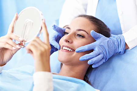 Woman receiving facial treatment with blue gloves on hands holding mirror.