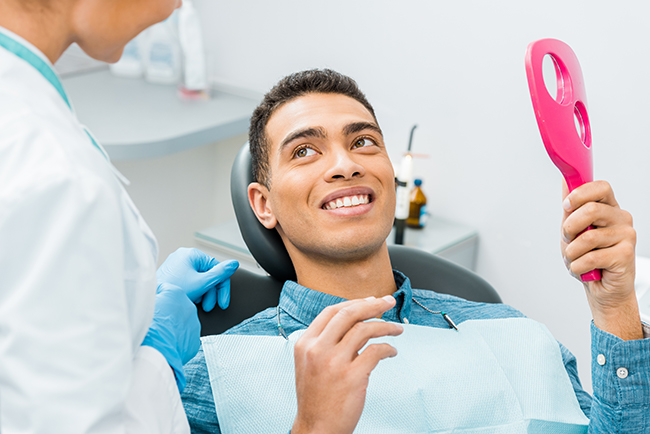 The image shows a man sitting in a dental chair with a smile on his face, holding a pink object, while being attended by a dental professional who appears to be performing a procedure.