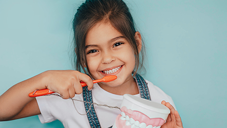 The image shows a young girl brushing her teeth with a red toothbrush while holding a toothpaste tube, smiling at the camera.
