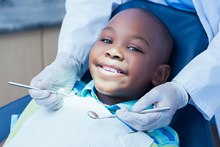 The image shows a young boy sitting in a dental chair with his mouth open, receiving dental care from a dentist wearing protective gloves.