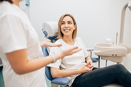 The image shows a woman sitting in a dental chair with a smile on her face, being attended by two dental professionals who are engaged in conversation with her.