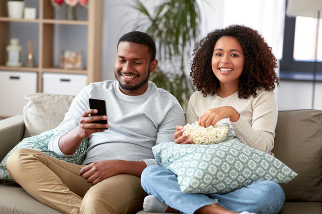 A man and woman sitting on a couch, sharing a moment of relaxation with a snack while watching something on their phones.