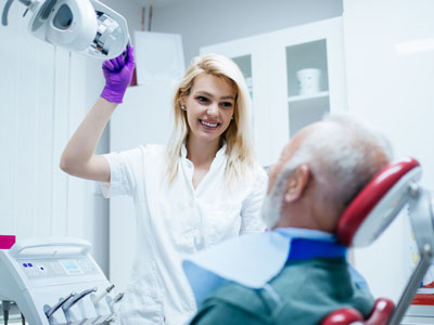 A dental hygienist with purple gloves and a white coat stands beside a patient who is seated in a chair, holding a mirror up to their face.