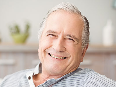 The image shows an elderly man with white hair smiling at the camera, wearing a blue shirt and seated comfortably in a living room setting.