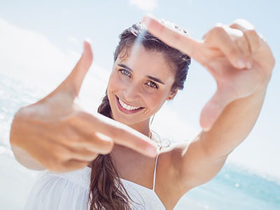 A woman is taking a selfie with her hand held up to the camera lens, smiling at the beach with clear skies and white sand in the background.
